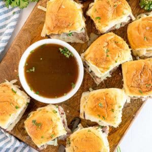 Sliders arranged around a bowl of au jus on a wooden board, viewed from above.