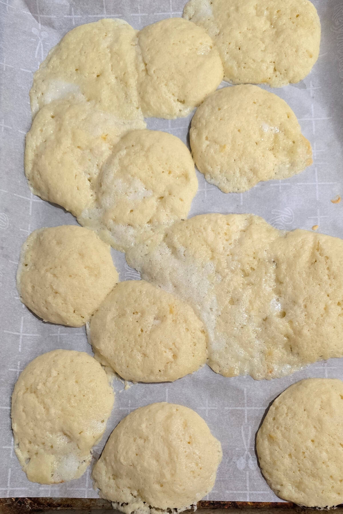 A baking tray lined with parchment paper holds unevenly shaped, pale yellow cookies, some of which have spread and merged together.