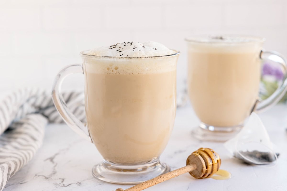 A tea latte with foam and sprinkled tea leaves in a glass mug. A honey dipper and tea bag beside it.
