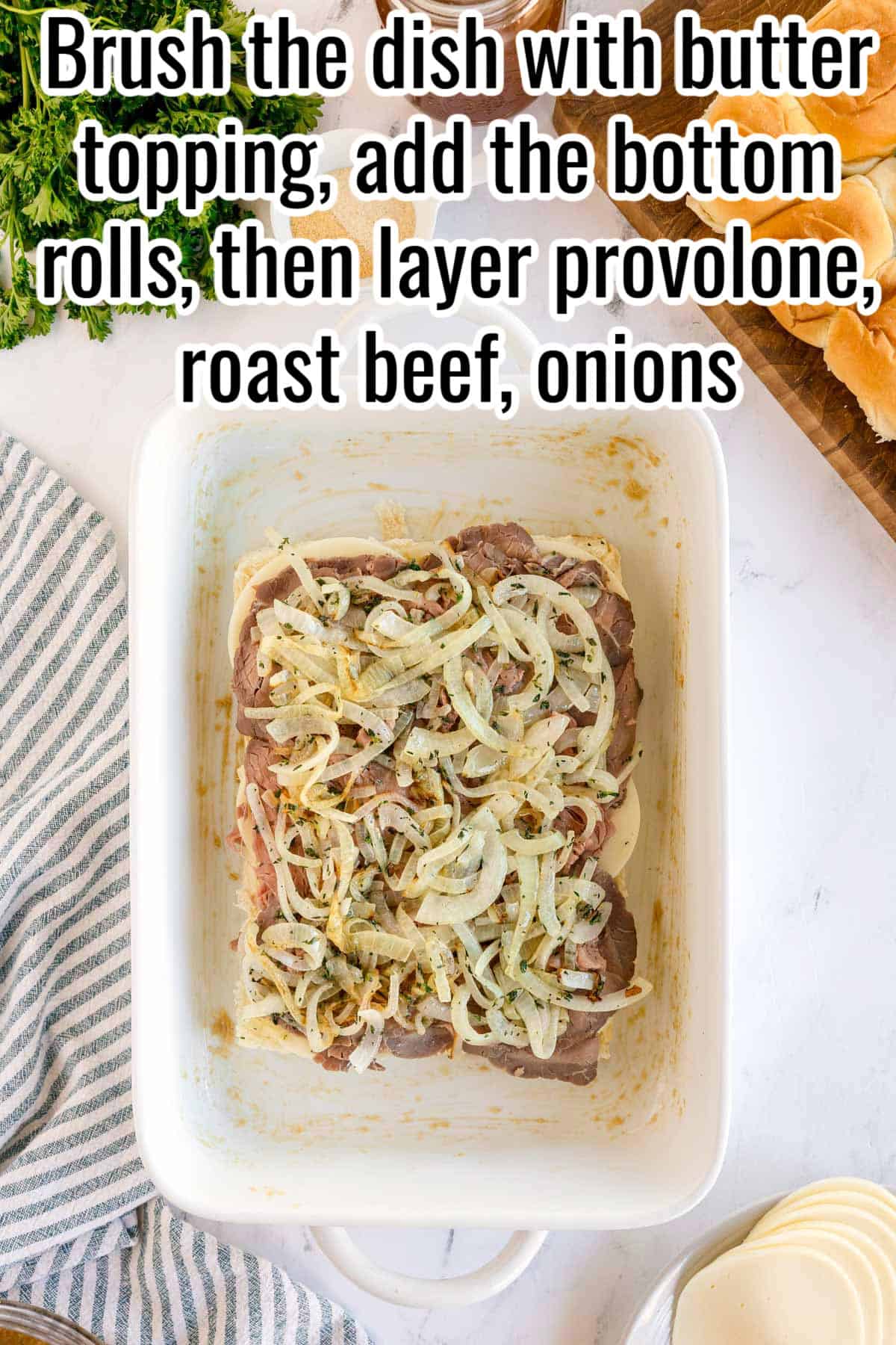 White baking dish with bottom slider buns layered with cheese, roast beef, and onions. Text on the image says 'Brush the dish with butter topping, add the bottom rolls, then layer provolone, roast beef, onions'.