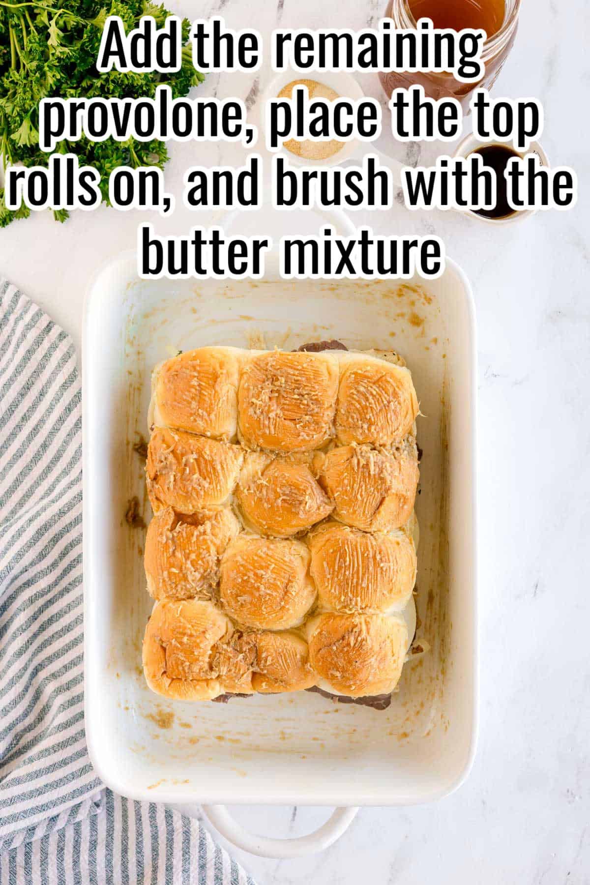 Baking dish with assembled sliders before baking. Text on the image says 'Add the remaining provolone, place the top rolls on, and brush with the butter mixture'.
