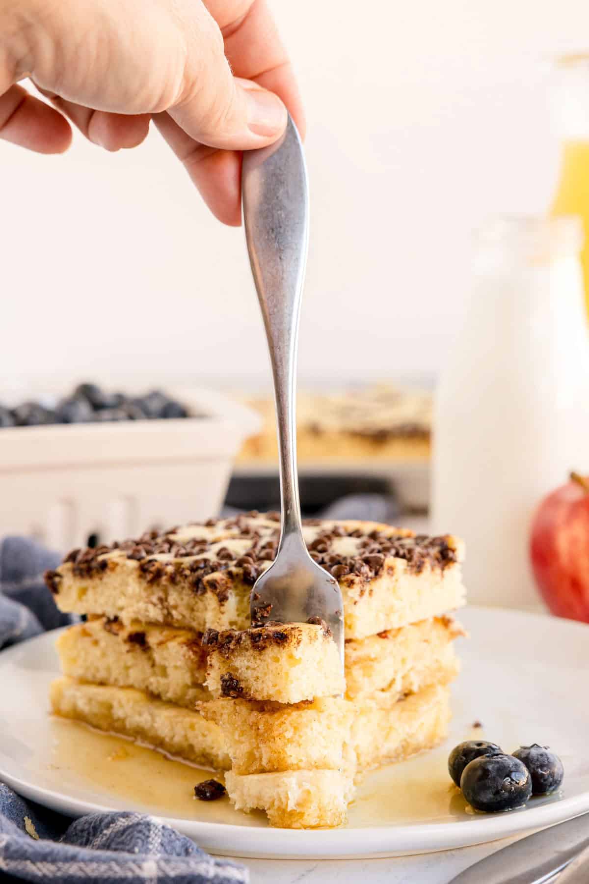 Fork cutting into a stack of chocolate chip-topped sheet pan pancakes with syrup and blueberries on the side.