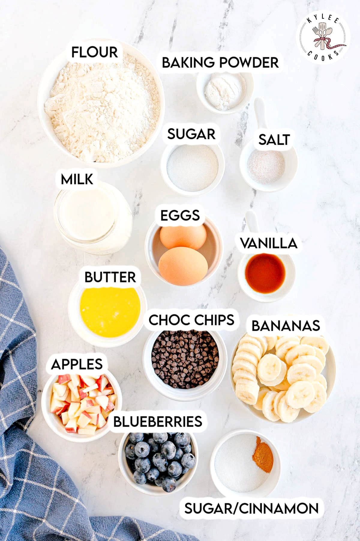 Bowls filled with labeled ingredients including flour, sugar, eggs, and fruit. Text on the image says 'FLOUR', 'BAKING POWDER', 'SUGAR', 'SALT', 'MILK', 'EGGS', 'VANILLA', 'BUTTER', 'CHOC CHIPS', 'BANANAS', 'APPLES', 'BLUEBERRIES', 'SUGAR/CINNAMON'.