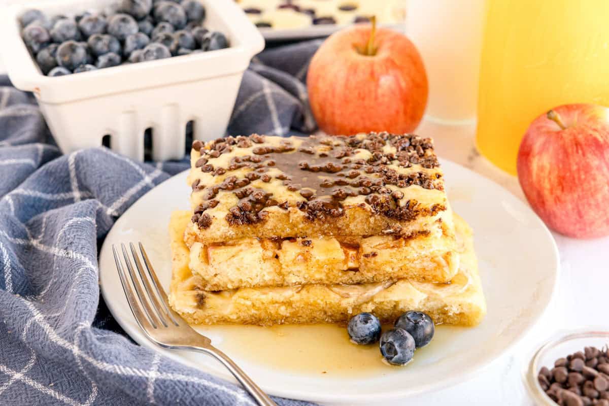 Stack of sheet pan pancakes topped with chocolate chips and syrup, with apples and blueberries on the side.