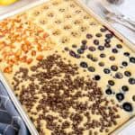Sheet pan pancake divided into sections topped with bananas, apples, blueberries, and chocolate chips.