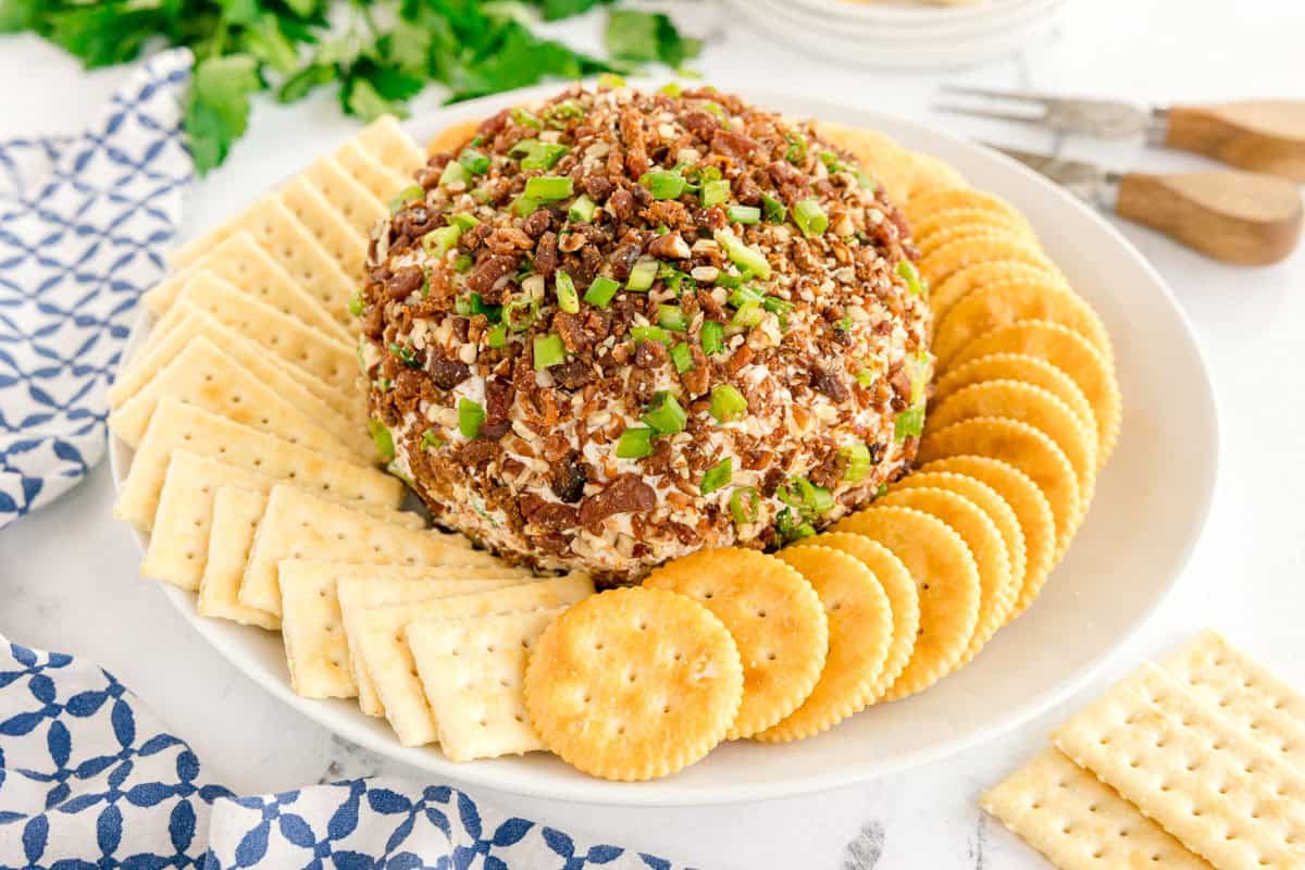 A whole bacon ranch cheese ball surrounded by square and round crackers on a white platter.
