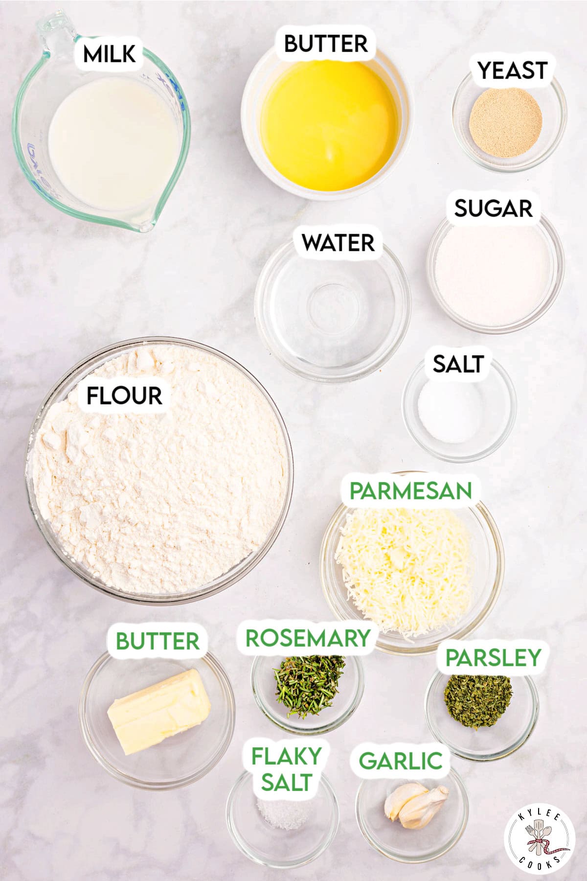 Ingredients in glass bowls labeled: flour, butter, milk, water, yeast, sugar, salt, parmesan, rosemary, parsley, flaky salt, and garlic. Text on the image says 'MILK', 'BUTTER', 'YEAST', 'SUGAR', 'WATER', 'SALT', 'FLOUR', 'BUTTER', 'ROSEMARY', 'FLAKY SALT', 'PARSLEY', 'GARLIC', 'PARMESAN'.