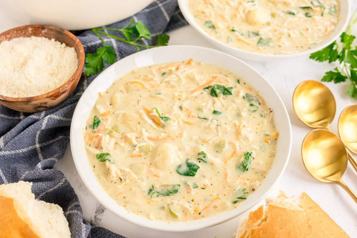 A bowl of creamy chicken gnocchi soup with spinach, shredded carrots, and celery. Parmesan and bread on the side.