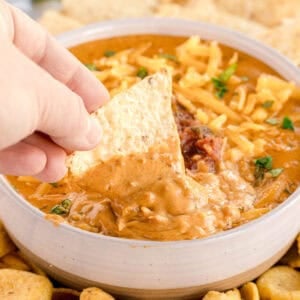 A hand dipping a tortilla chip into cheesy chili dip in a bowl.