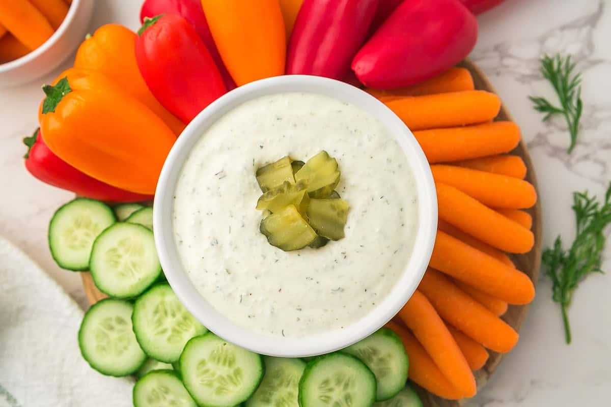 A bowl of creamy Dill Pickle Dip, made with cottage cheese and topped with chopped pickles, is surrounded by sliced cucumbers, baby carrots, and red and orange mini bell peppers.