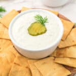 A bowl of dip topped with a pickle slice and dill, surrounded by pita chips on a round board.