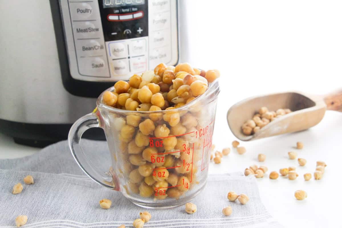 A glass measuring cup filled with cooked chickpeas, with an Instant Pot and scoop of dried chickpeas nearby.