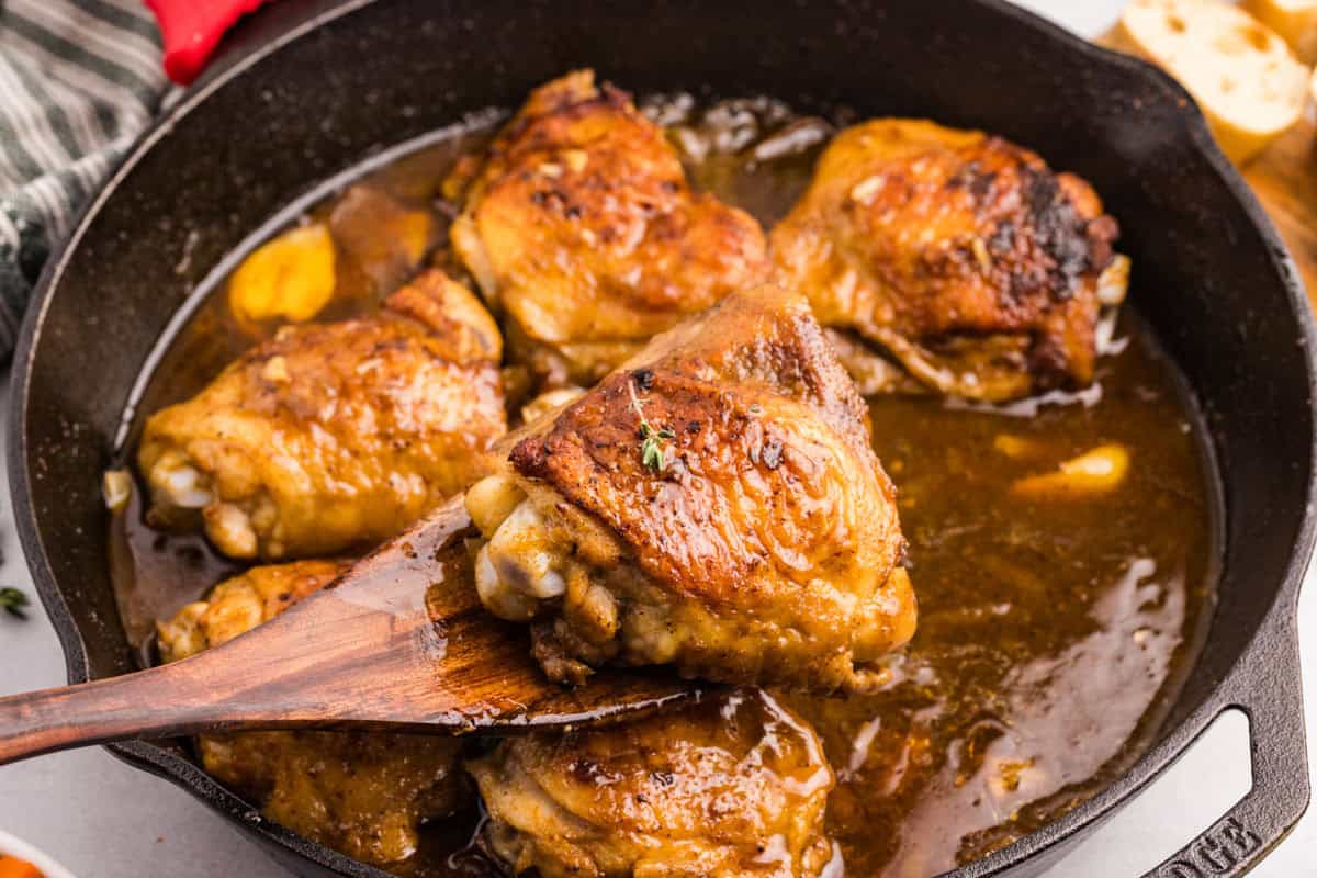 A skillet with browned chicken thighs in a golden sauce, one lifted on a wooden spatula.