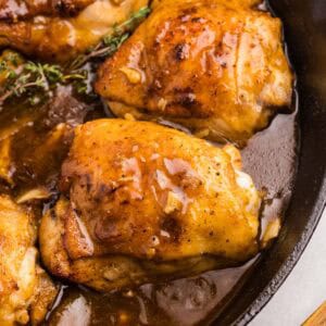 Close-up of chicken thighs in a skillet with rich sauce and herbs.