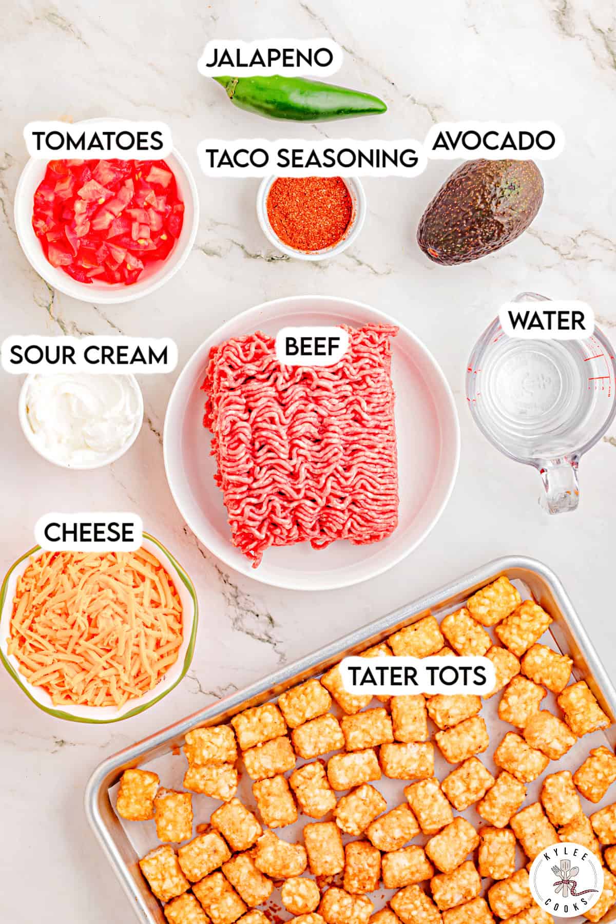 Bowls and trays of ingredients labeled as tomatoes, taco seasoning, avocado, jalapeño, sour cream, beef, cheese, water, and tater tots.