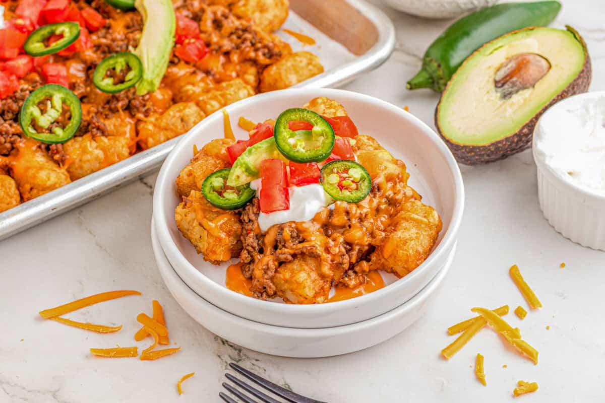 A small bowl of totchos with beef, cheese, tomatoes, avocado, jalapeños, and sour cream, next to a tray of totchos.