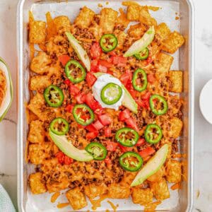 A square view of totchos on a tray, topped with beef, cheese, tomatoes, avocado slices, jalapeños, and sour cream.