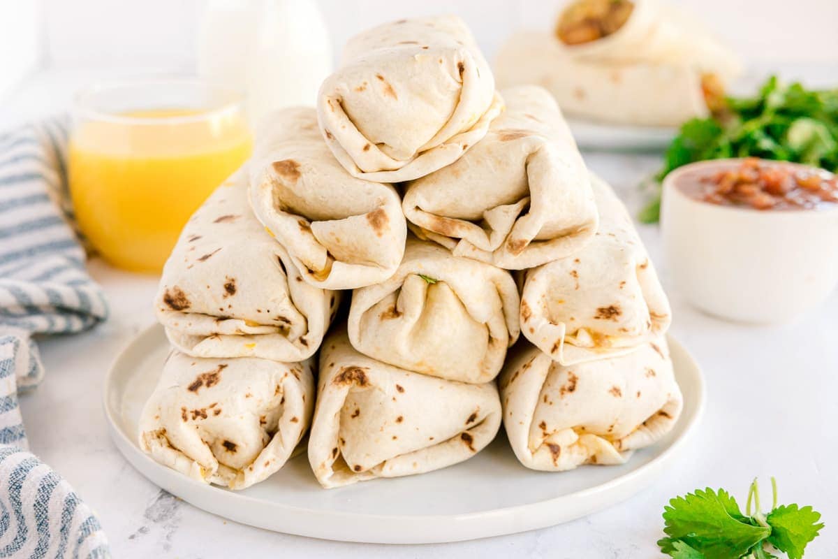 Stack of wrapped breakfast burritos on a plate with a glass of orange juice in the background.