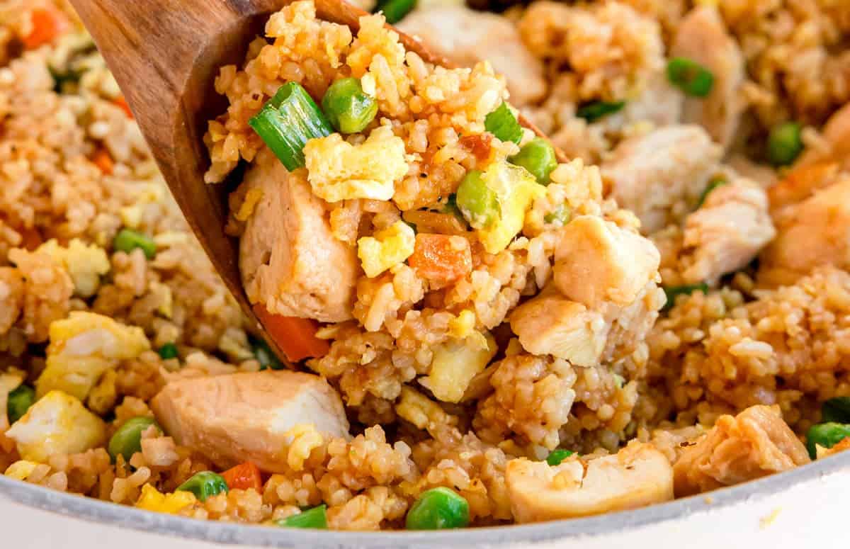 Wooden spoon scooping chicken fried rice with peas, carrots, eggs, and green onions from a skillet.