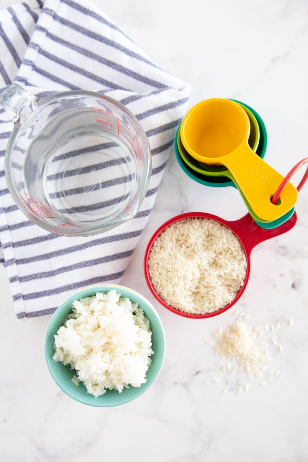 A measuring cup of water, a bowl of cooked rice, a cup of uncooked rice, measuring spoons, and a striped towel on a white surface—perfect essentials for any rice guide or to help you cook rice with ease.