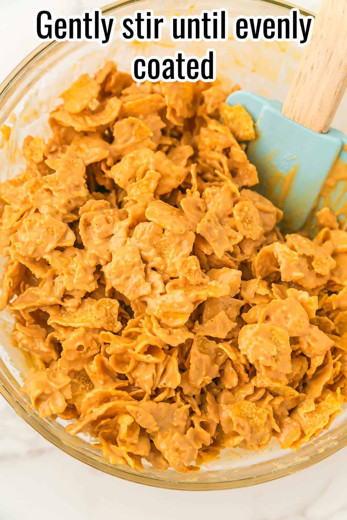 Cornflakes coated in peanut butter mixture with a spatula in the bowl. Text on the image says 'Gently stir until evenly coated'.