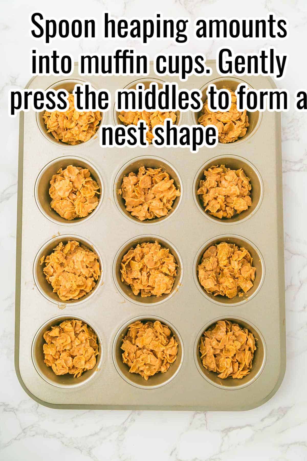 Cornflake mixture pressed into muffin tin cups. Text on the image says 'Spoon heaping amounts into muffin cups. Gently press the middles to form a nest shape'.