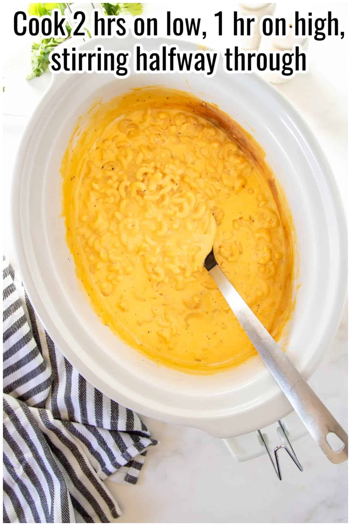 Creamy macaroni and cheese in a slow cooker with a spoon inside. Text on the image says 'Cook 2 hrs on low, 1 hr on high, stirring halfway through'.