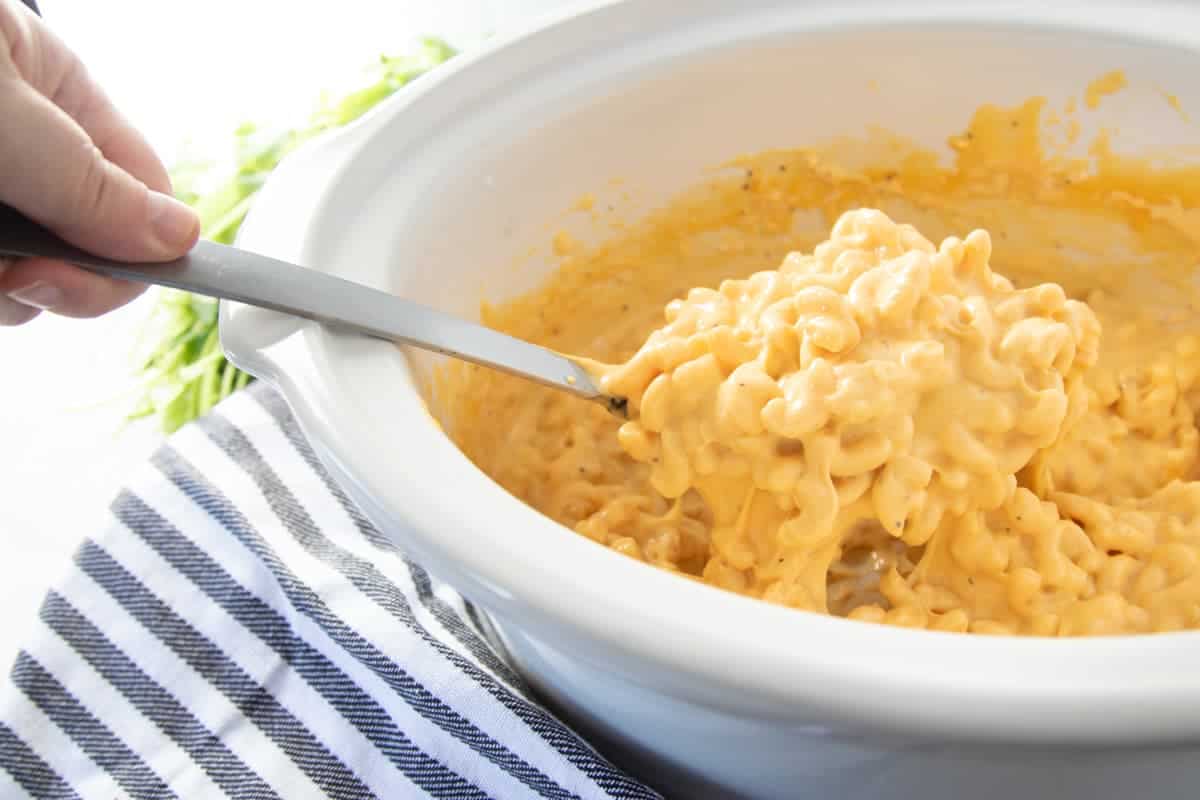 Spoon lifting creamy Velveeta mac and cheese from a white slow cooker.