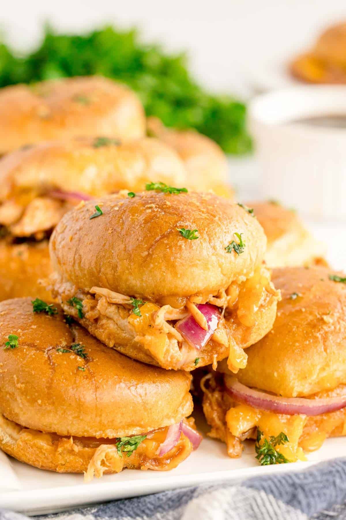 A close-up of BBQ chicken sliders with red onions and melted cheese on golden buns, garnished with chopped parsley, arranged on a white plate.