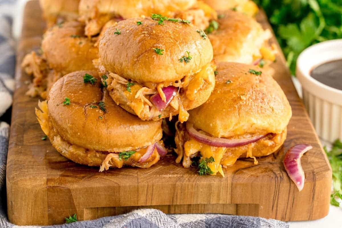 A wooden board with pulled chicken sliders, cheddar cheese, and red onion on buns, garnished with chopped parsley. A small dish of barbecue sauce is visible in the background.