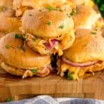 Close-up of several pulled chicken sliders with melted cheese and red onion on toasted buns, garnished with chopped parsley, arranged on a wooden board.