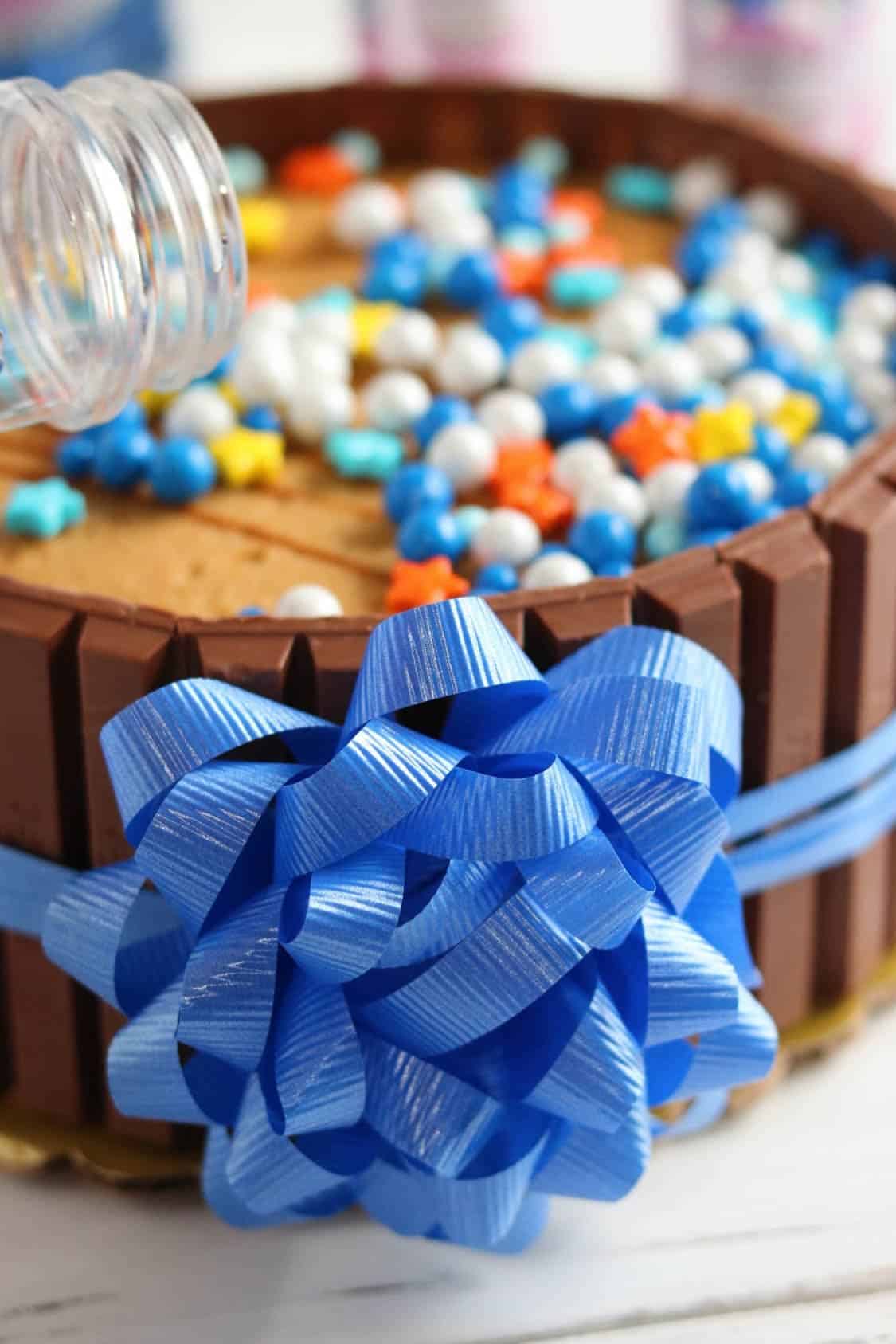 Pouring blue and white candy sprinkles onto a KitKat cake with a blue ribbon bow.