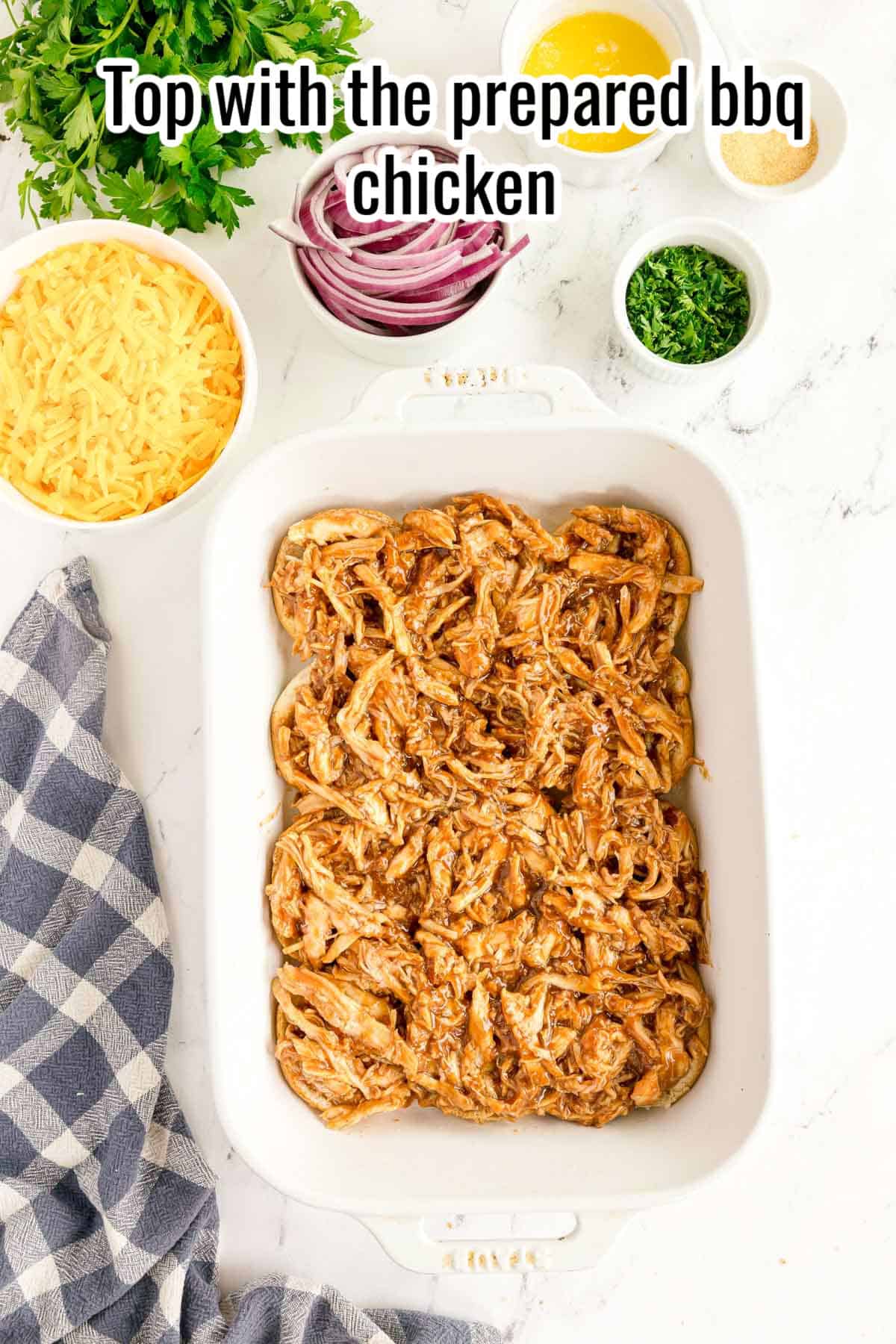 A baking dish filled with shredded BBQ chicken sits on a counter surrounded by bowls of shredded cheese, parsley, red onion, and a checked towel—perfect for assembling delicious bbq chicken sliders. Instructions read: "Top with the prepared bbq chicken.