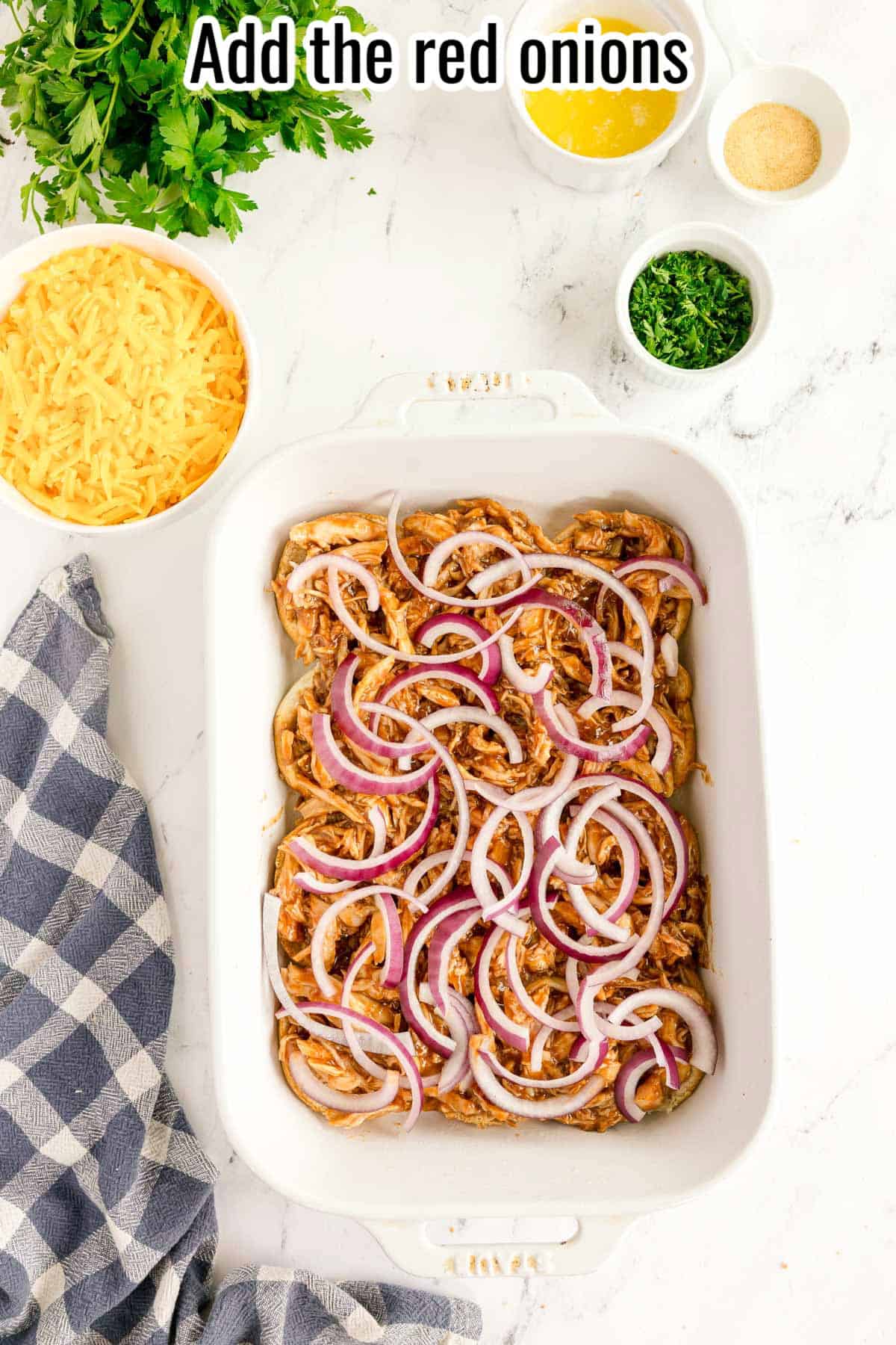 A white baking dish filled with shredded bbq chicken sliders topped with sliced red onions sits on a marble counter, surrounded by cheese, herbs, and a blue checkered towel. Text at top reads, "Add the red onions.