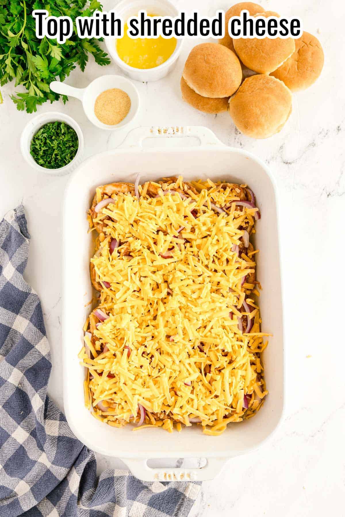 A white baking dish filled with shredded cheese over layered ingredients for bbq chicken sliders, surrounded by buns, cheese sauce, parsley, and seasonings on a white countertop. Text reads: "Top with shredded cheese.