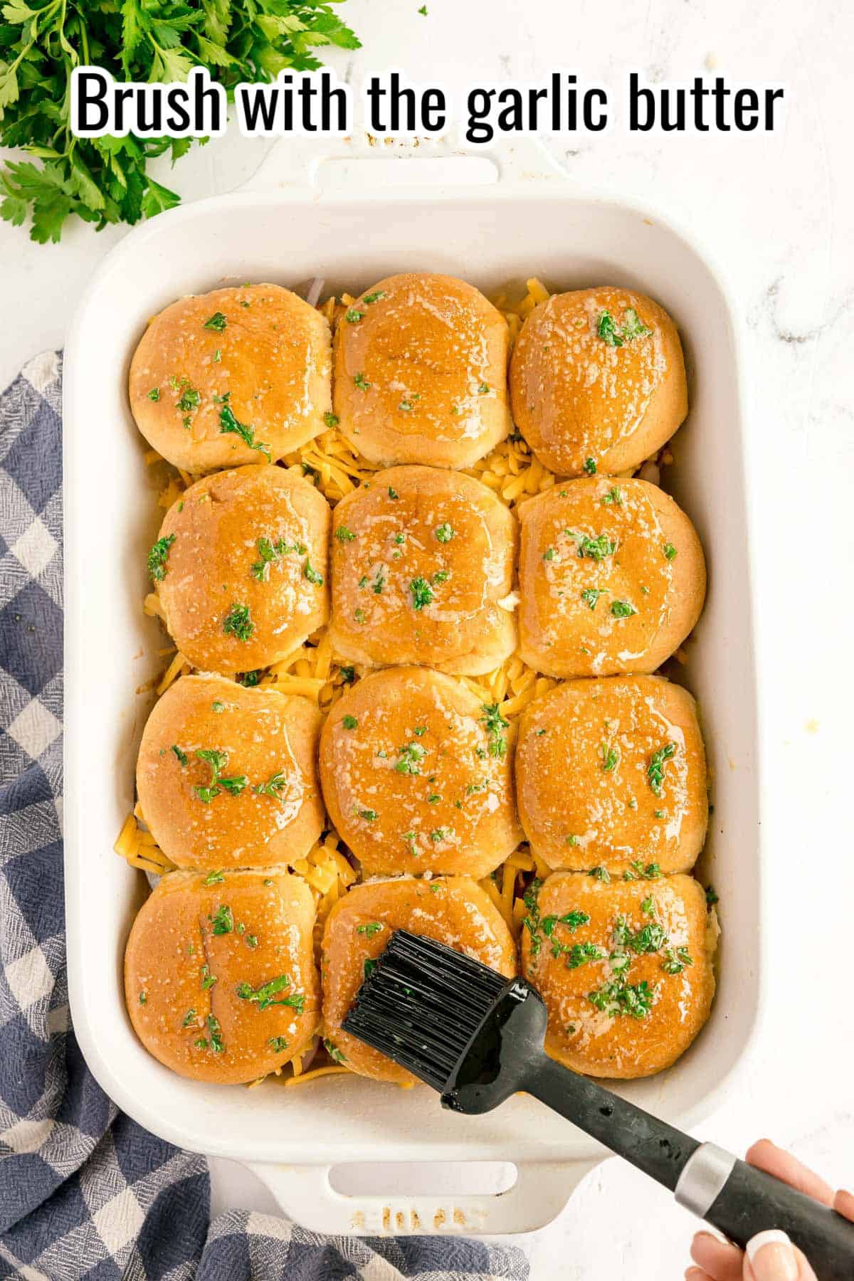 A hand brushes garlic butter on top of twelve bbq chicken sliders in a white baking dish, with chopped parsley and a checked towel nearby.