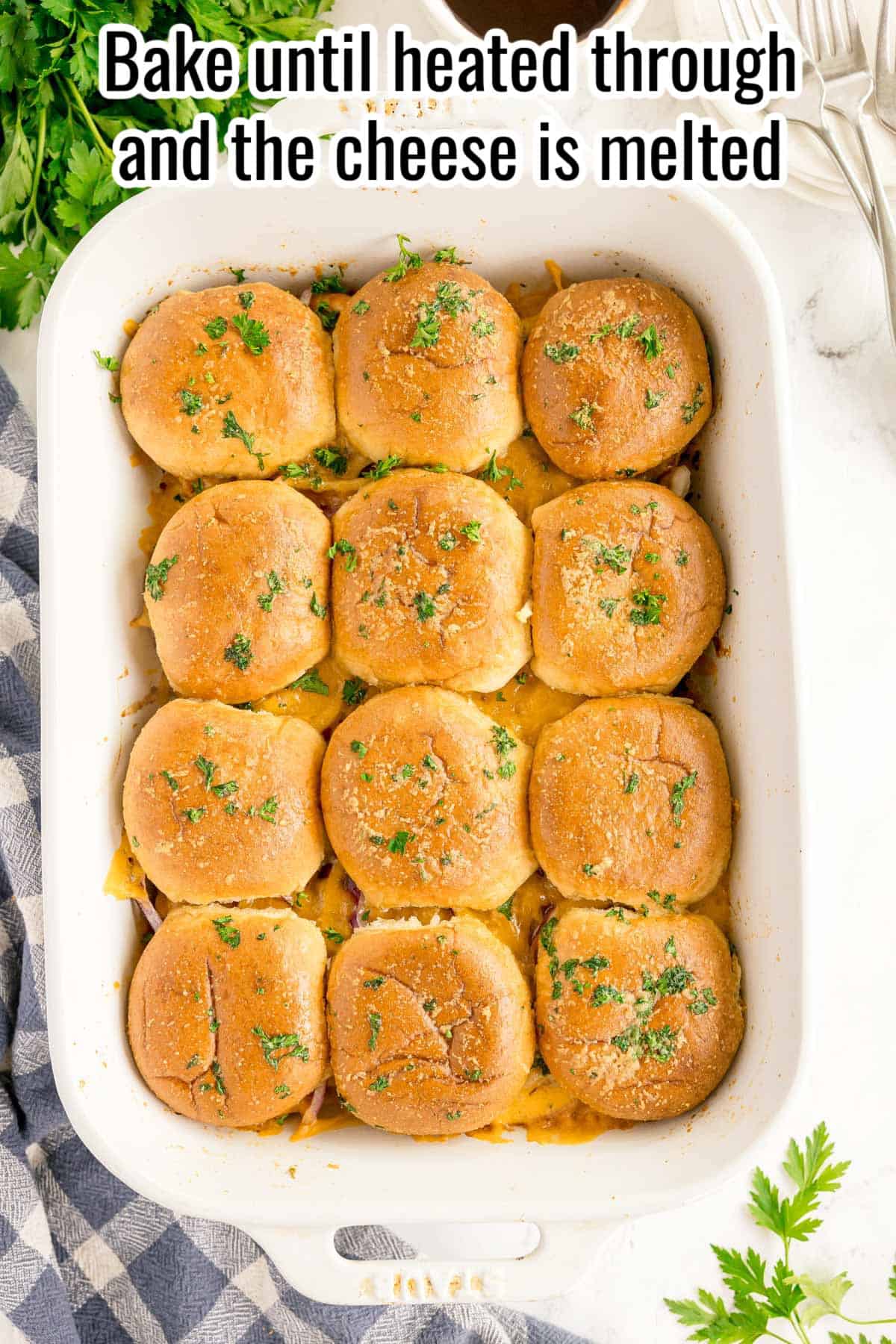 A white baking dish with twelve baked BBQ chicken sliders topped with melted cheese and chopped parsley, next to a blue plaid napkin and fresh herbs. Text at top reads, "Bake until heated through and the cheese is melted.