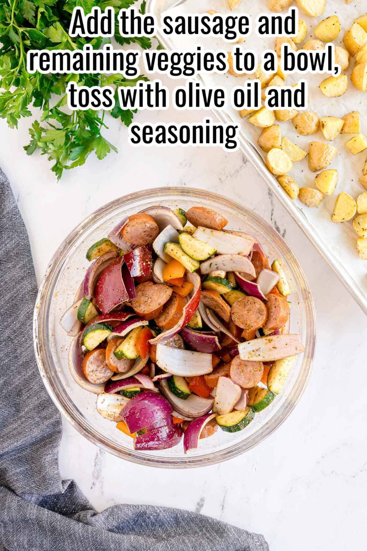Step two of making sheet pan sausage and veggies: sliced sausage, zucchini, red onion, and bell peppers tossed together in a large glass bowl, with the partially roasted potatoes visible on the sheet pan in the background. Text reads: Add the sausage and remaining veggies to a bowl, toss with olive oil and seasoning.