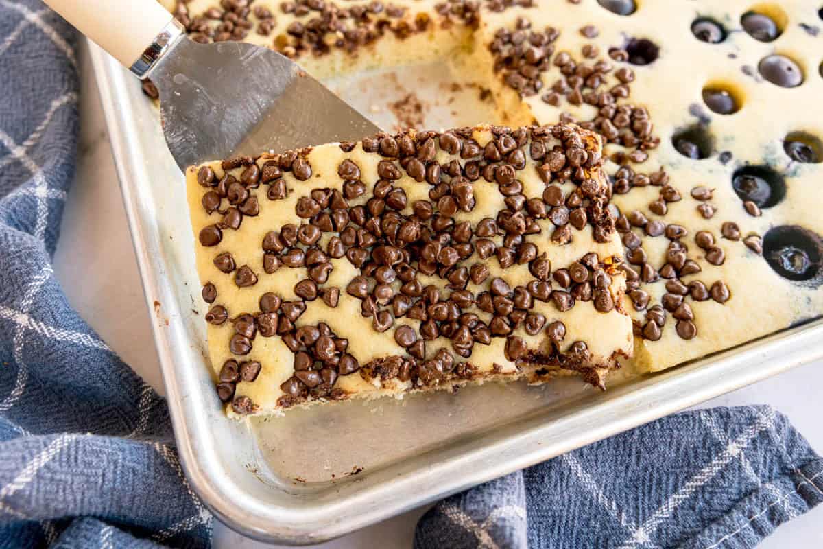 A sheet pan filled with baked pancake topped with chocolate chips, with one large square piece being lifted ou.