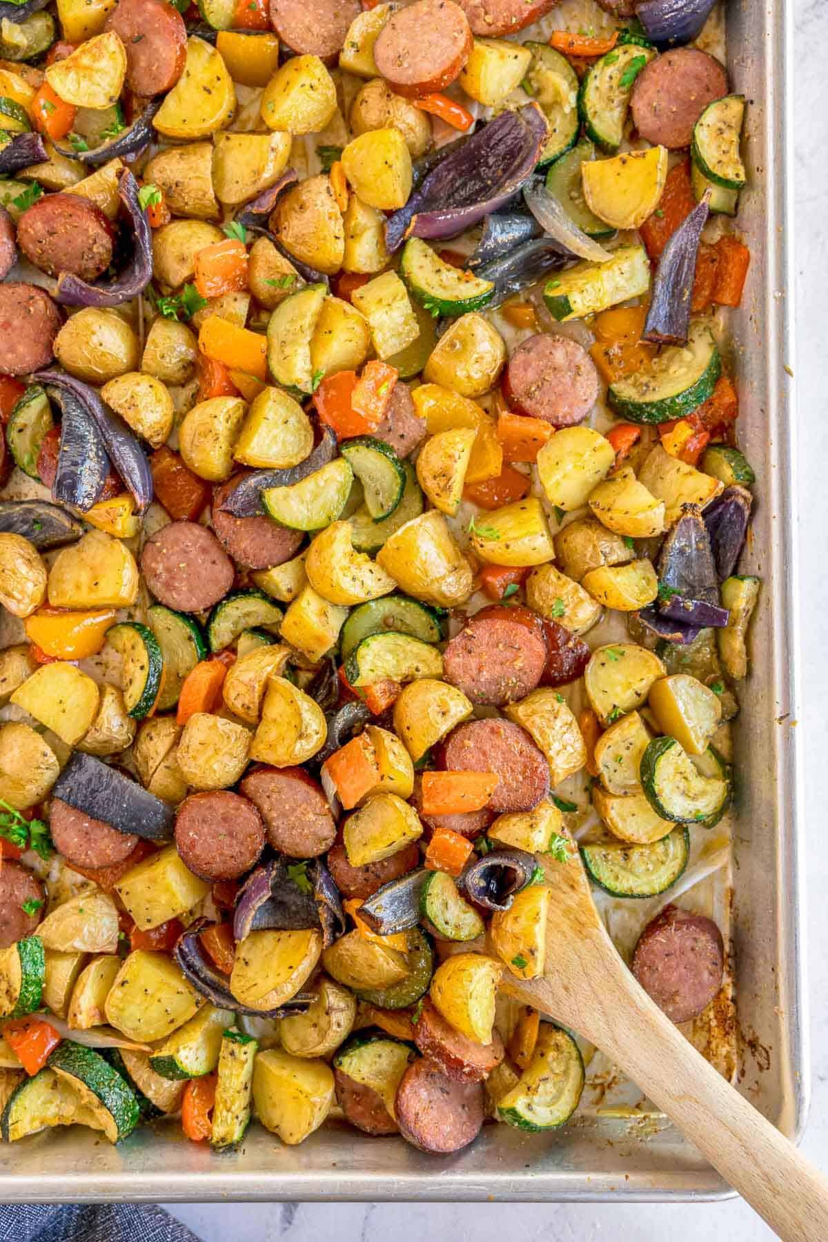 Overhead close-up of a sheet pan of roasted sausage and vegetables including potatoes, zucchini, red onion, and bell peppers, with a wooden spoon resting in the pan.