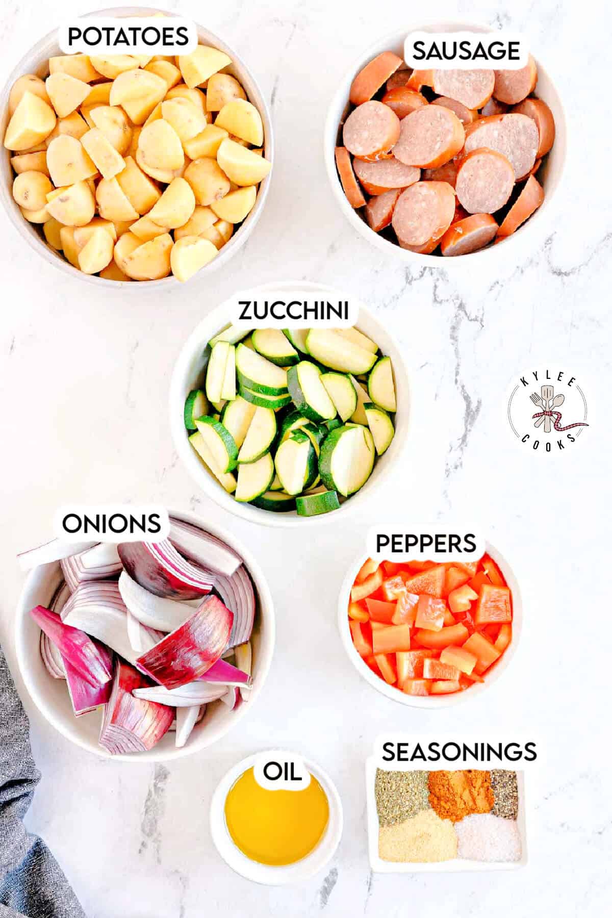 Flat lay of sheet pan sausage and veggies ingredients in white bowls on a marble surface, including potatoes, sliced sausage, zucchini, red onion wedges, diced peppers, oil, and seasonings.