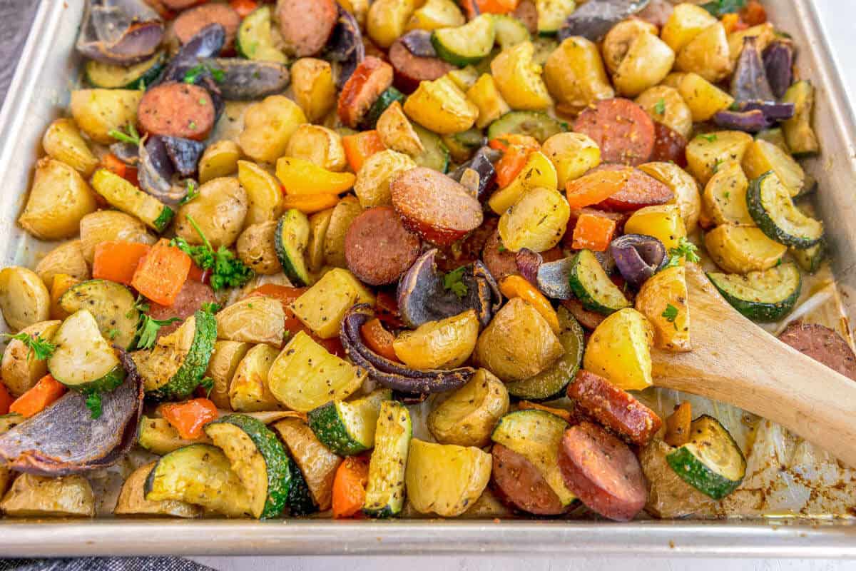 A close-up of a sheet pan filled with roasted sausage slices, potatoes, zucchini, red onion, bell peppers, and carrots, garnished with fresh parsley, with a wooden spoon resting in the pan.