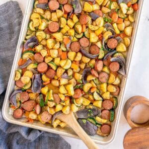 Overhead view of a sheet pan filled with roasted sausage, potatoes, zucchini, red onion, and bell peppers, garnished with fresh parsley, with a wooden spoon and salt and pepper shakers alongside a gray linen.