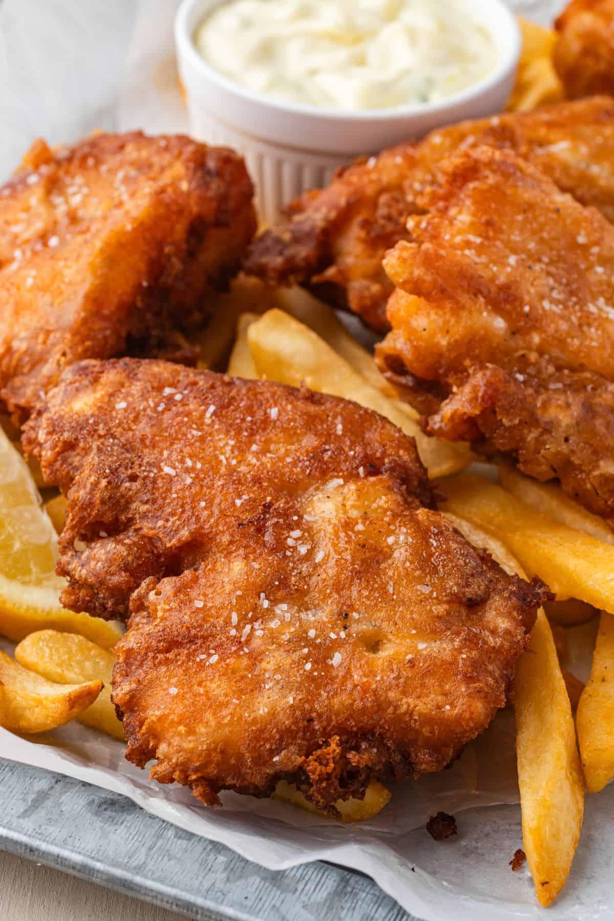 A plate of golden-brown fried fish, thick-cut fries, and lemon wedges, served with a small cup of tartar sauce in the background.