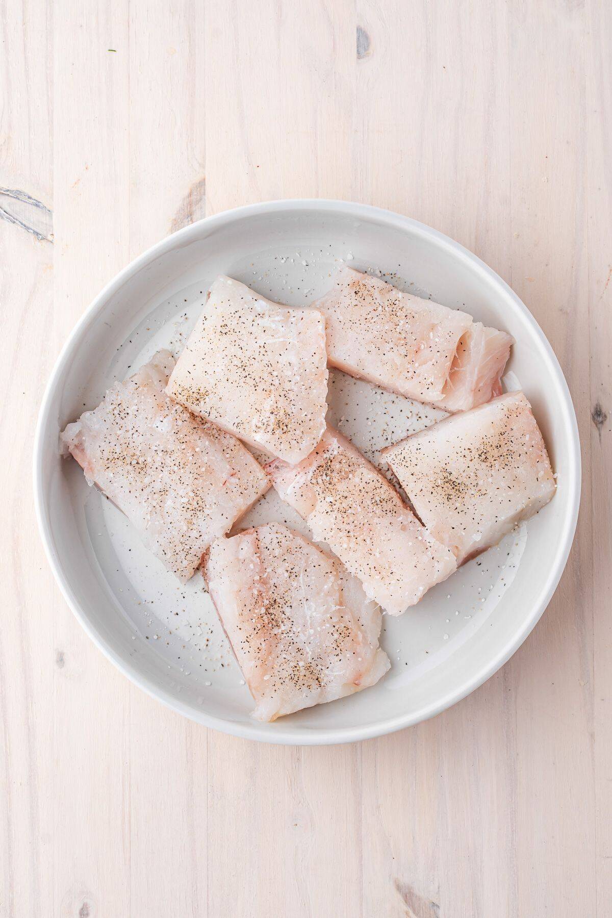 A white dish contains six raw fish fillets seasoned with salt and pepper, placed on a light wooden surface.