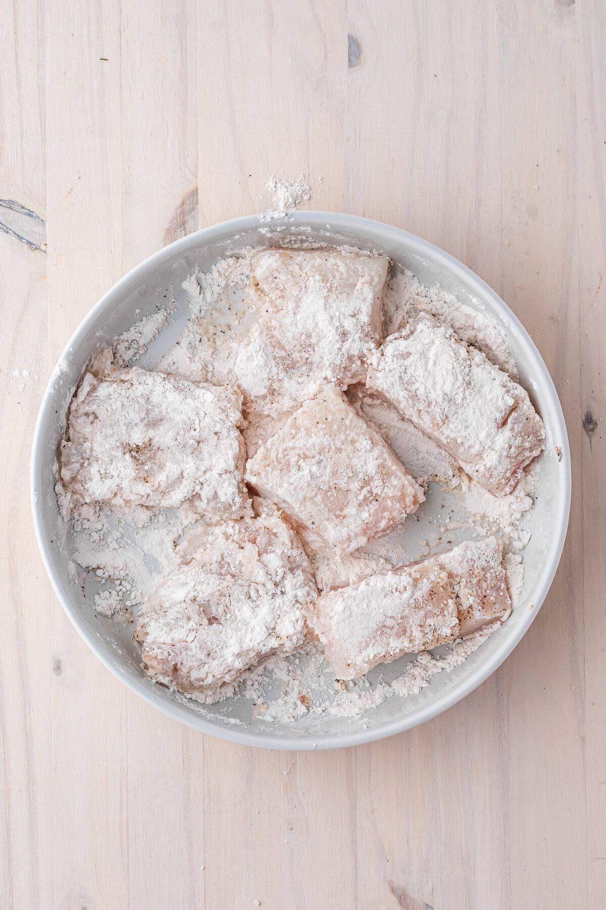 A white plate with several pieces of raw fish coated in flour on a light wooden surface.