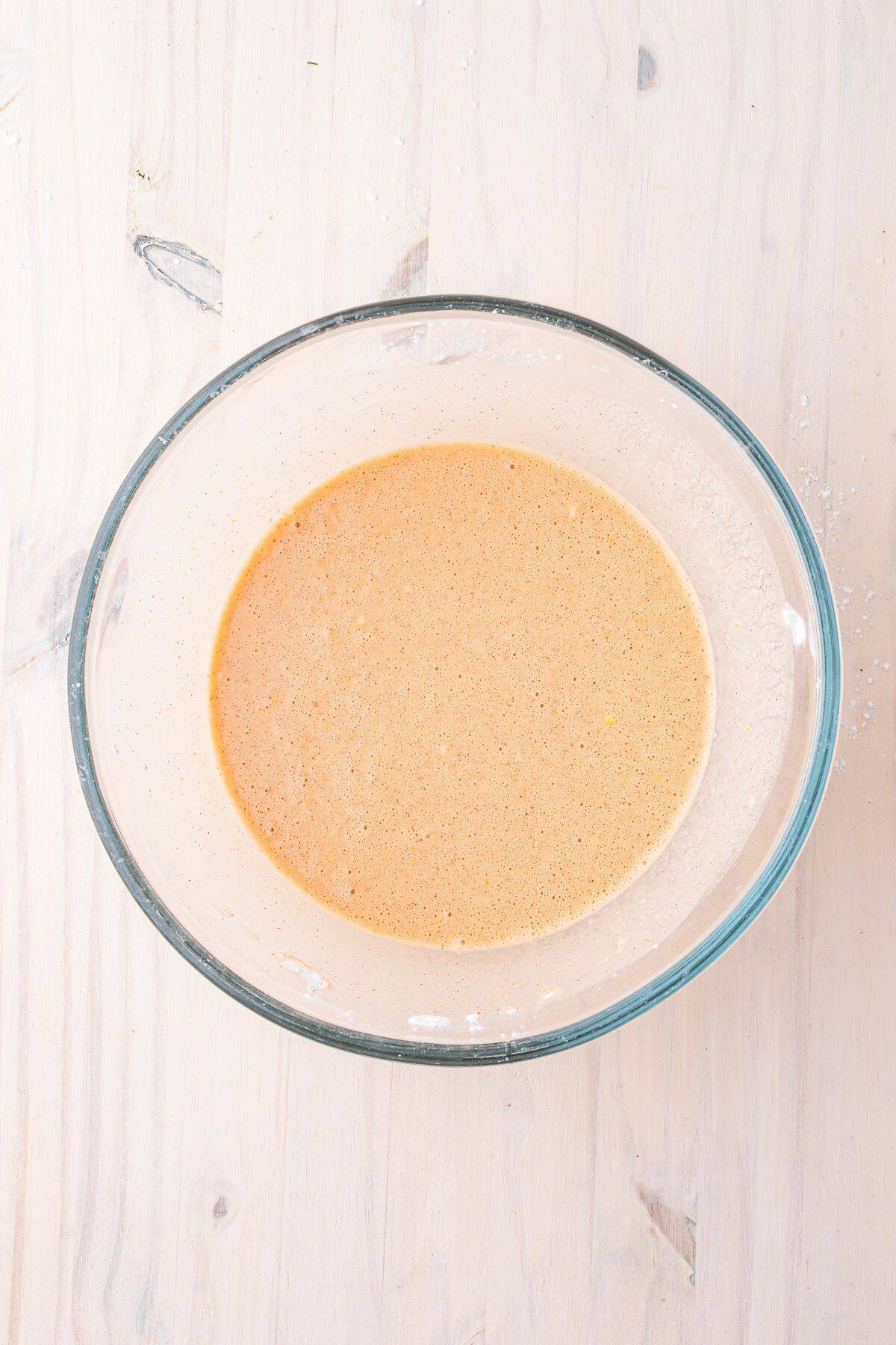 A glass bowl filled with a light brown liquid mixture sits on a light wooden surface.
