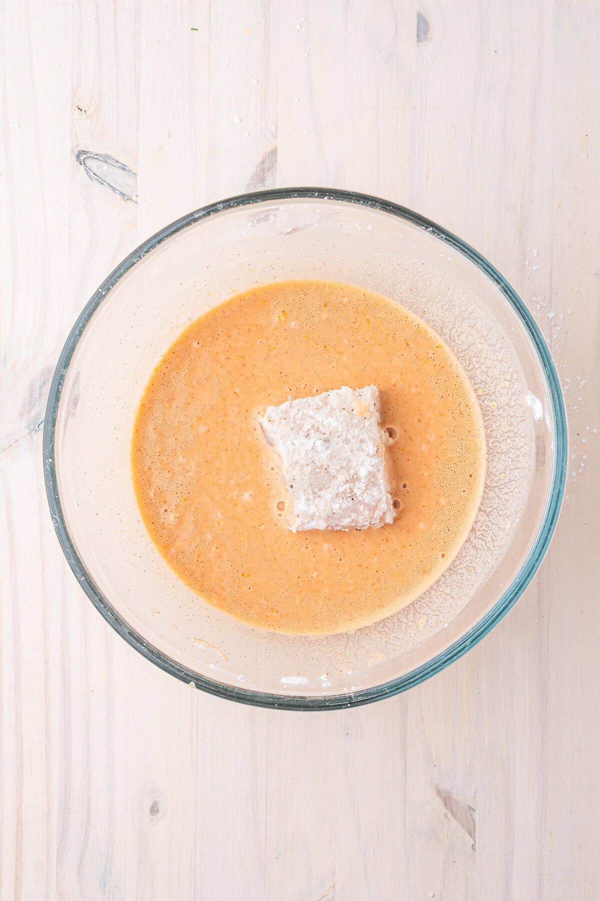 A piece of fish coated in flour is placed in an orange-colored batter inside a clear glass bowl on a light wooden surface.