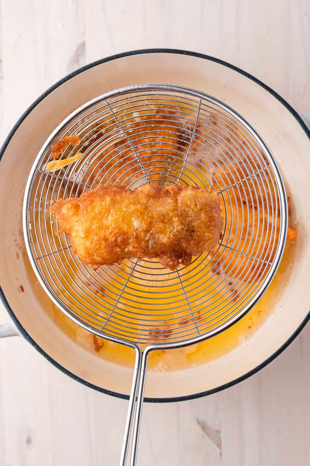 A piece of battered fish being lifted from a pot of hot oil with a metal skimmer.