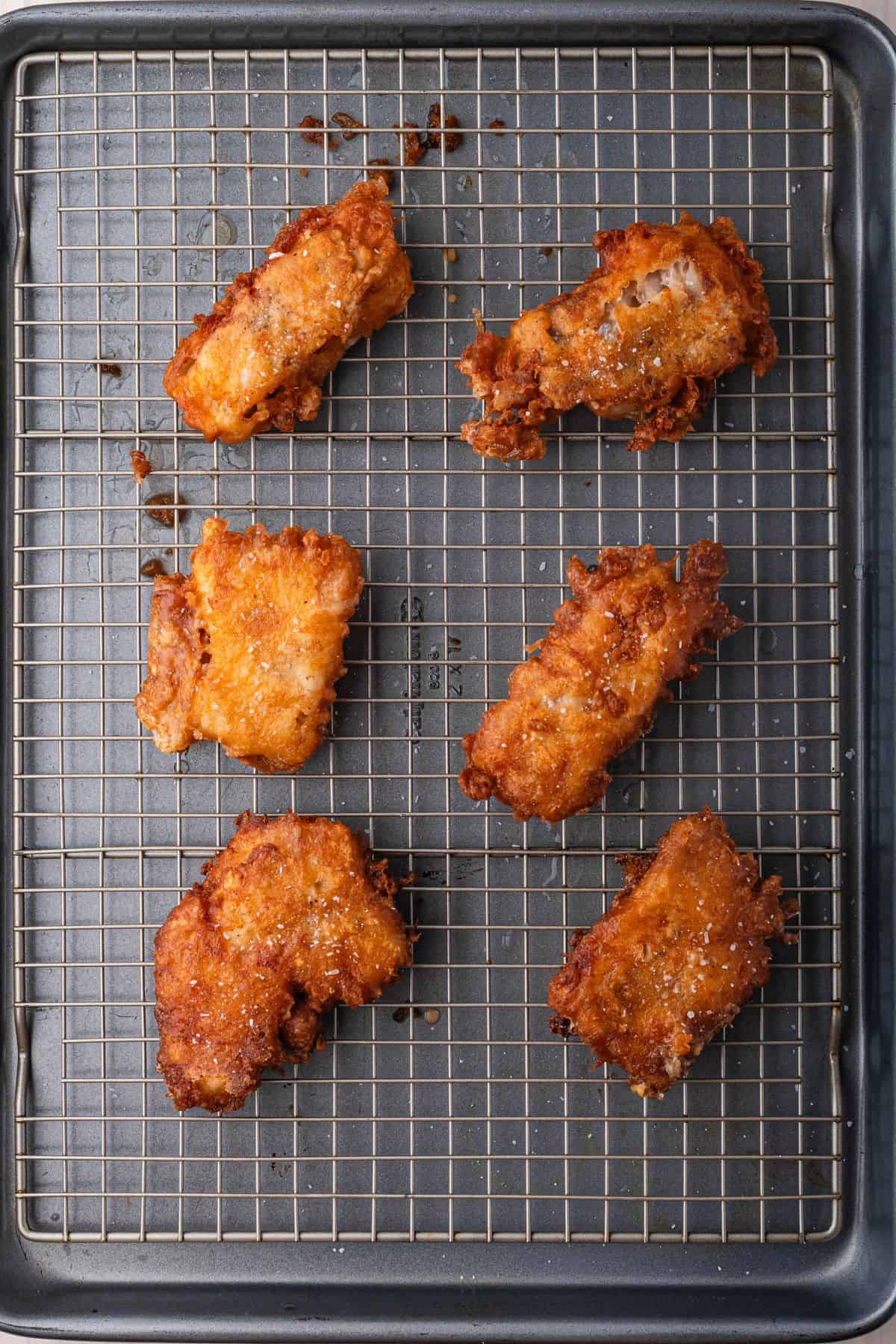 Six pieces of golden-brown fried chicken placed on a wire rack over a baking sheet.